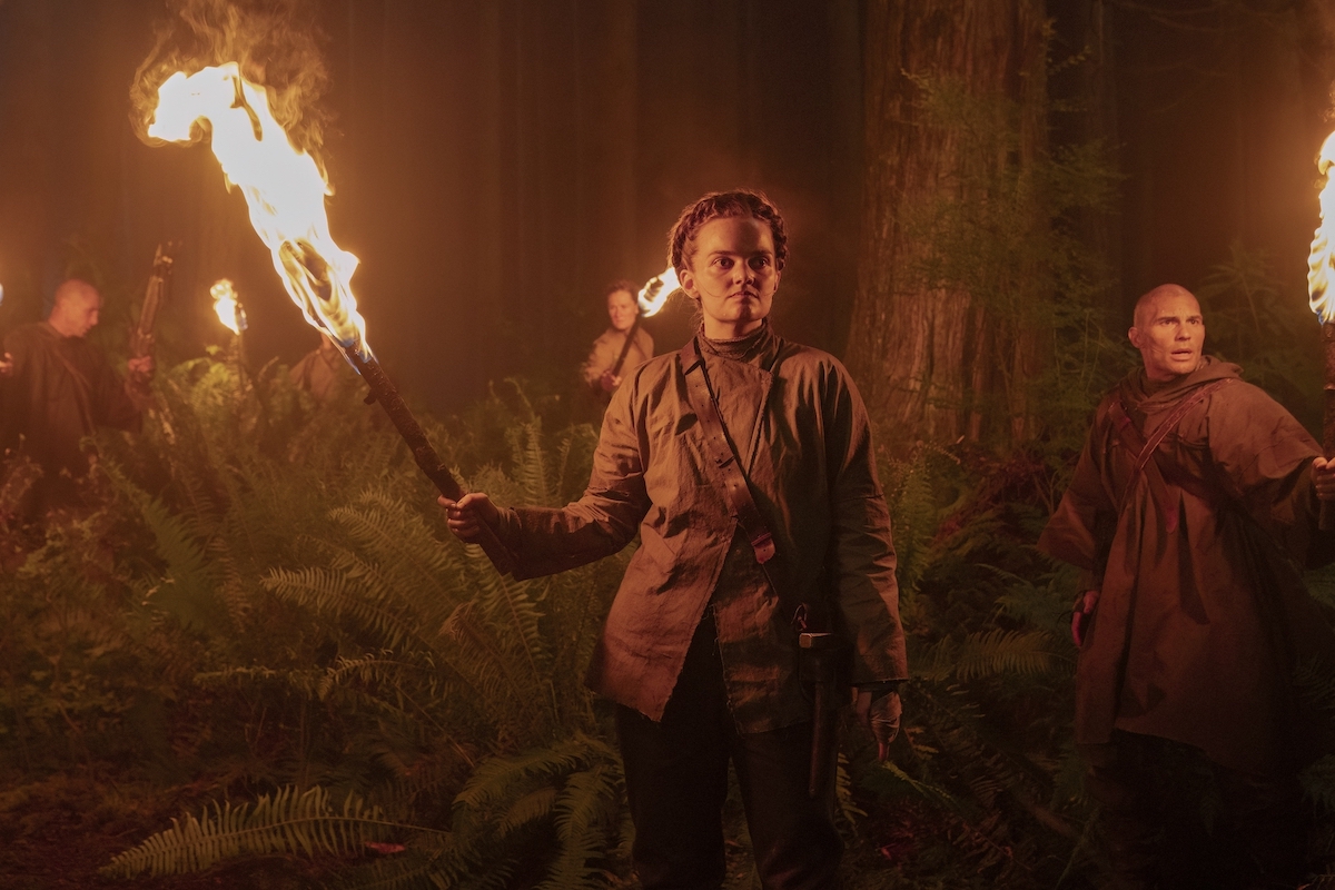 Scar / Seraphite member Laine MacNeil in 'The Last of Us' Season 2 Episode 5, shown holding a torch in the woods with other scars / seraphites