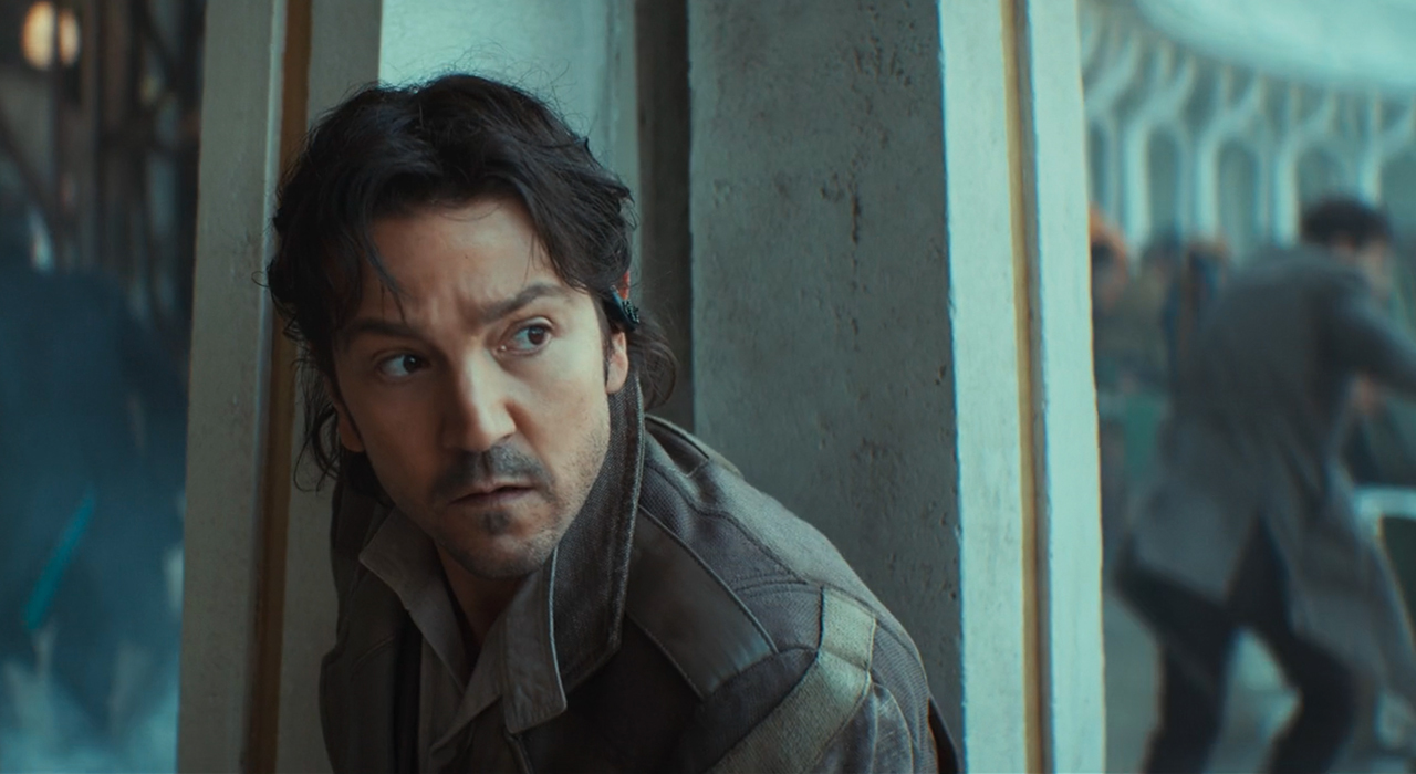 Cassian Andor (Diego Luna) hiding behind a stone column with a choatic crowd scene out of focus behind him in Episode 8 of Season 2 of 'Andor'
