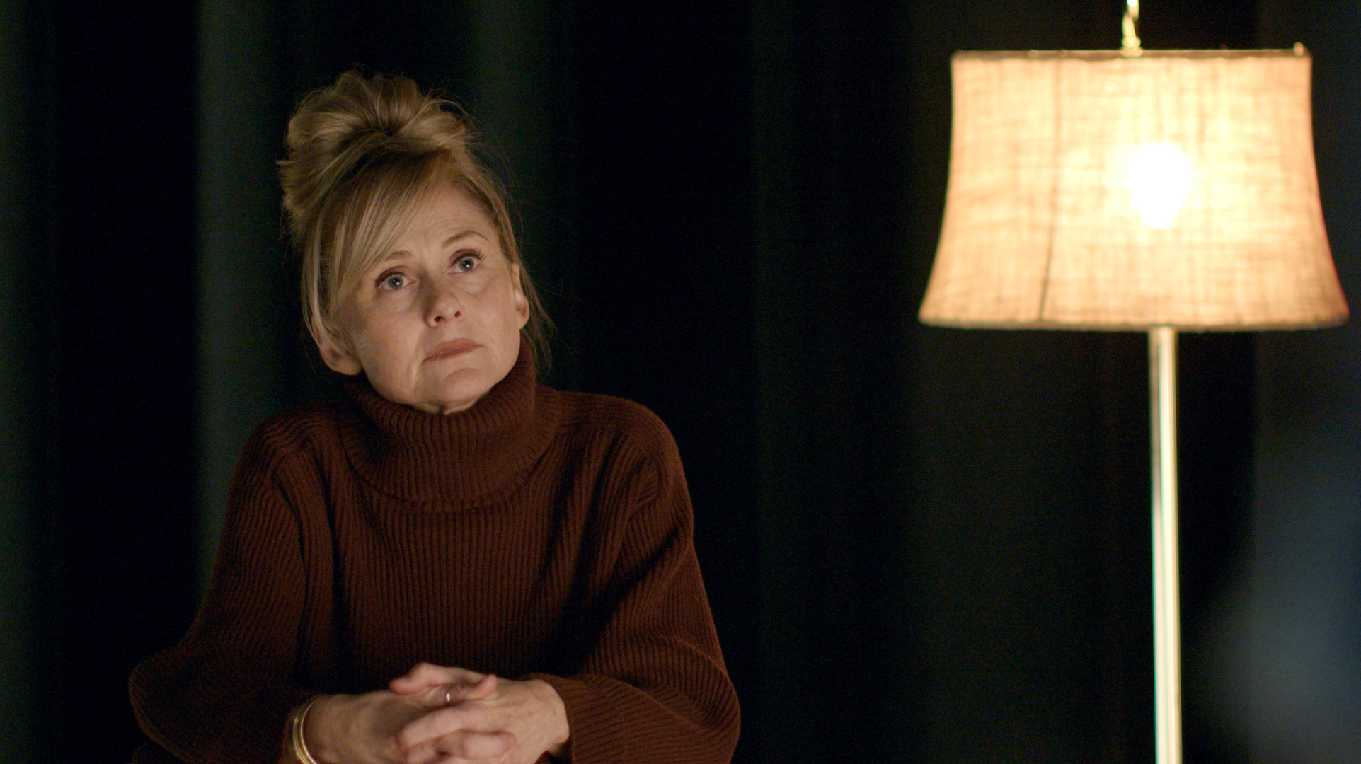 A woman in a maroon sweater sitting near a standing lamp; Heather Kafka in 'Leads'