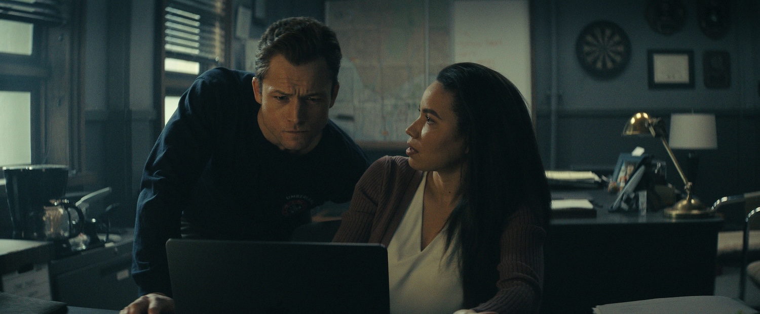 Taron Egerton and Jurnee Smollett in 'Smoke,' shown here with Taron looking over her shoulder at the computer, and Jurnee looking accusingly at him