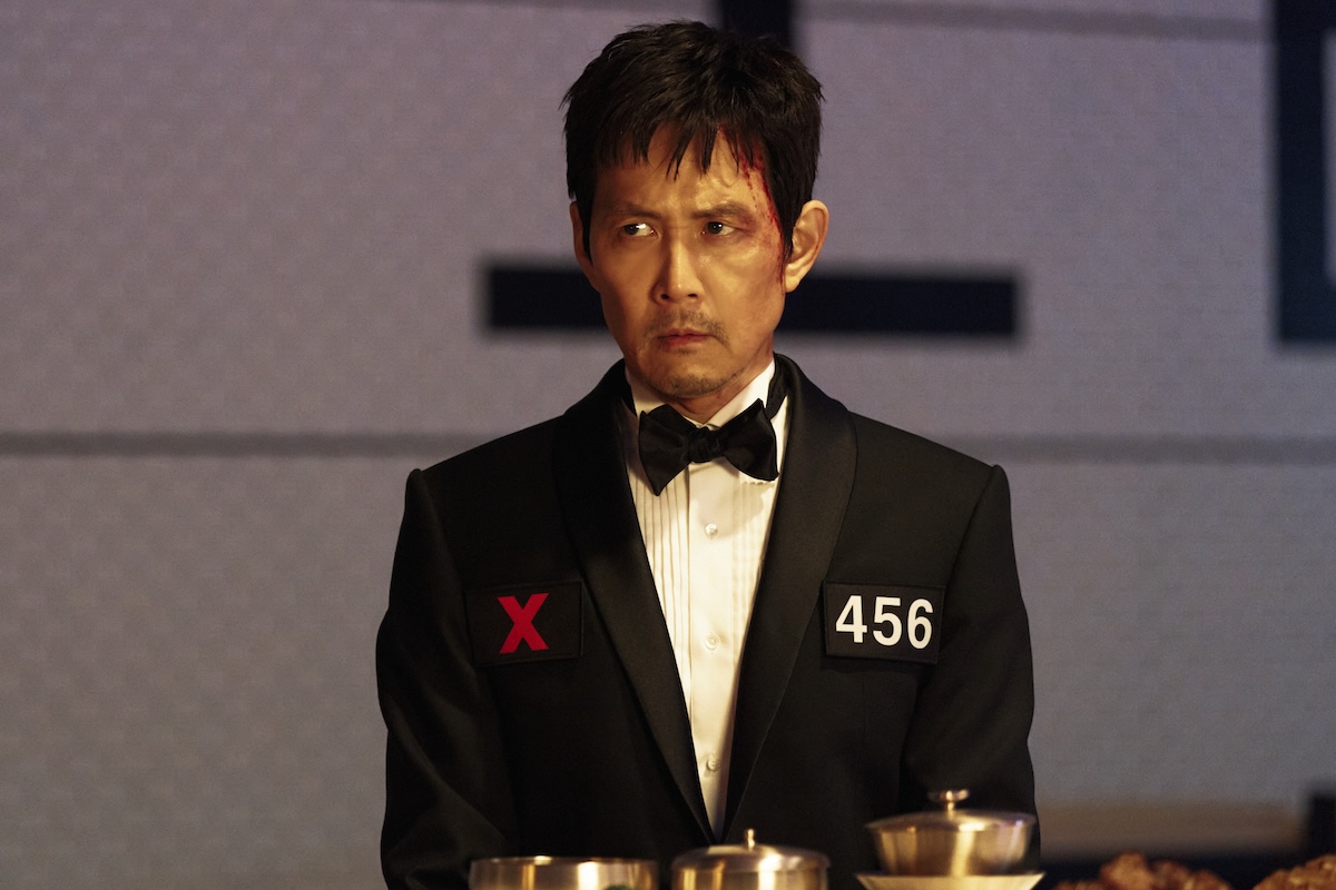 Squid Game Season 3 stars Lee Jung-jae as Seong Gi-hun, shown here with blood dripping down his face, wearing a tuxedo with 456 and a red X on it