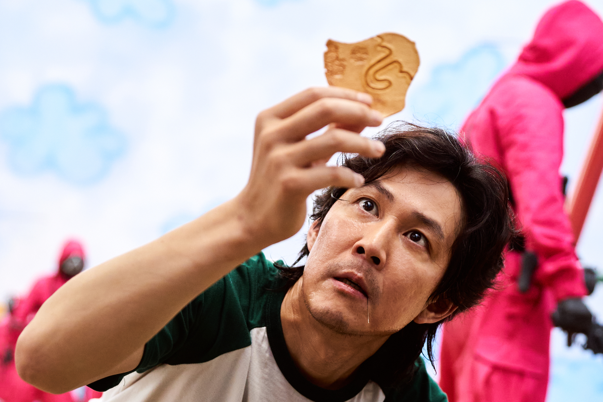 Close up of a man intensely examining a cookie while hooded guards in red walk around behind him; still from "Squid Game."