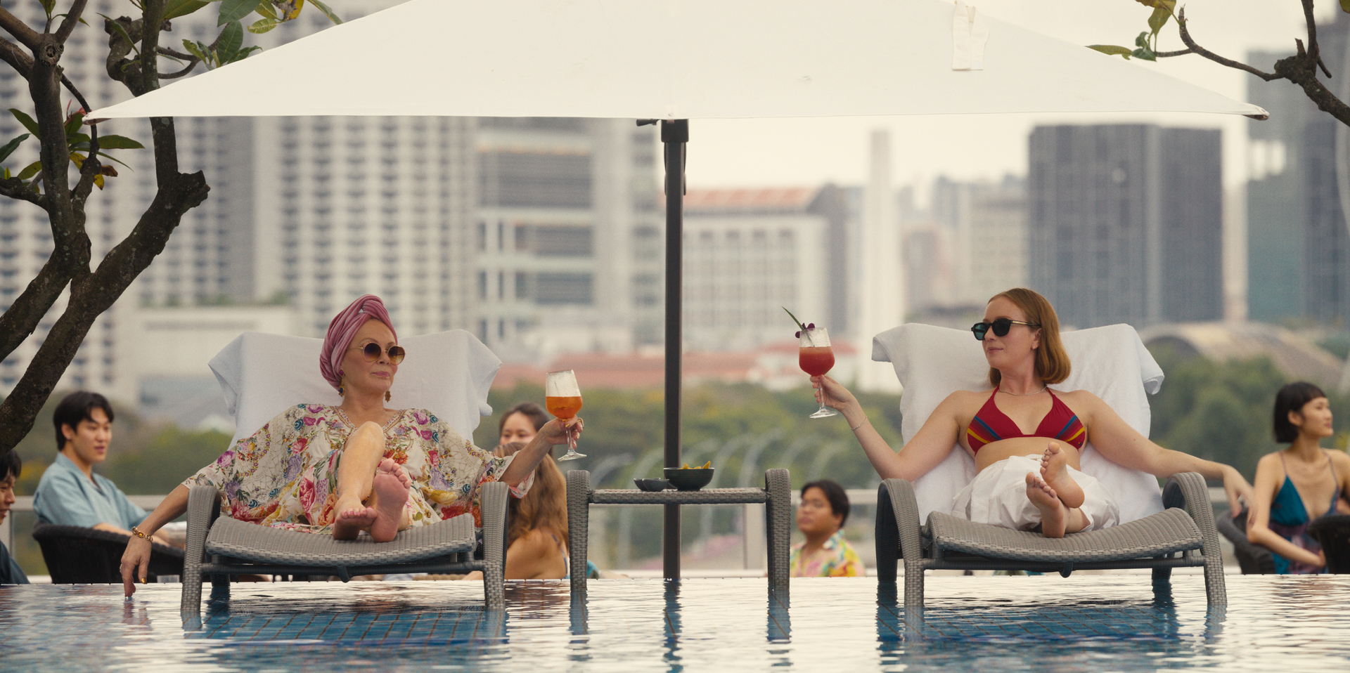 Jean Smart and Hannah Einbinder as Deborah and Ava in Season 4 of Hacks, longing on pool chairs with the Singapore skyline behind them.