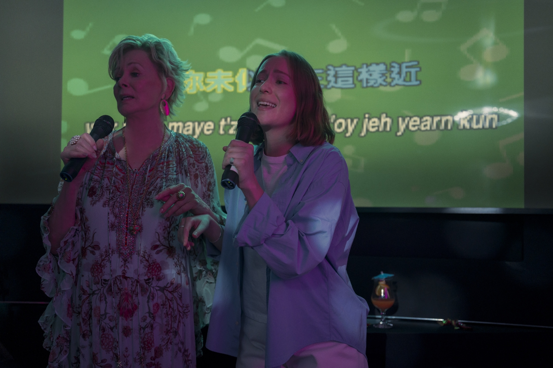 Jean Smart and Hannah Einbinder as Deborah and Ava in Season 4 of Hacks, doing Karaoke with mics in their hands and a screen showing Mandarin lyrics to The Cranberries 'Dreams' behind them. 