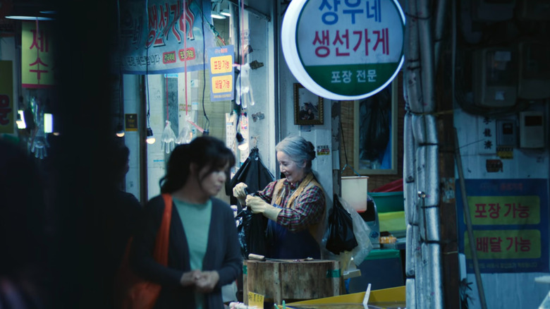 Old woman next to a sign of Sang-woo's fish market on Squid Game