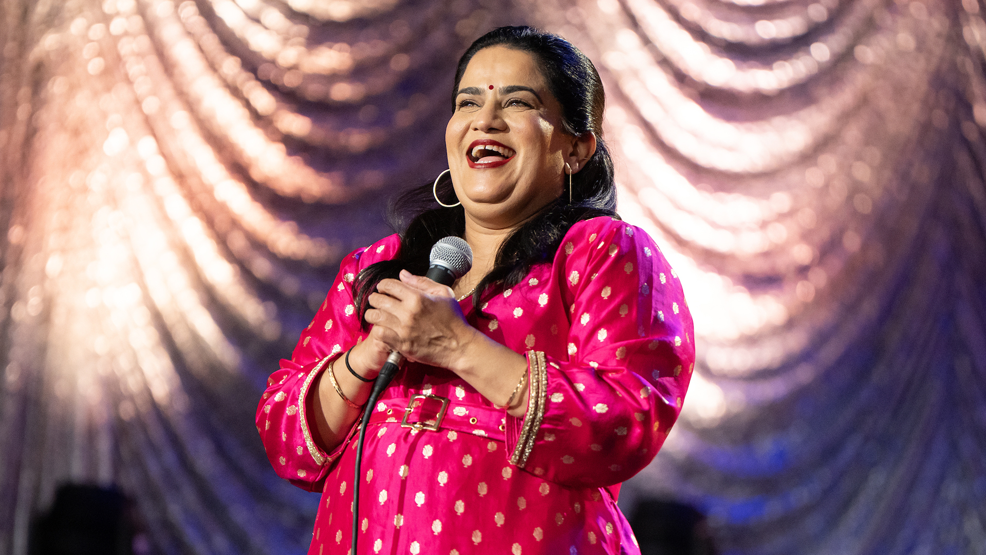 Zarna Garg wearing a bright pink kurta and laughing while holding a mic on stage in 'Practical People Win'