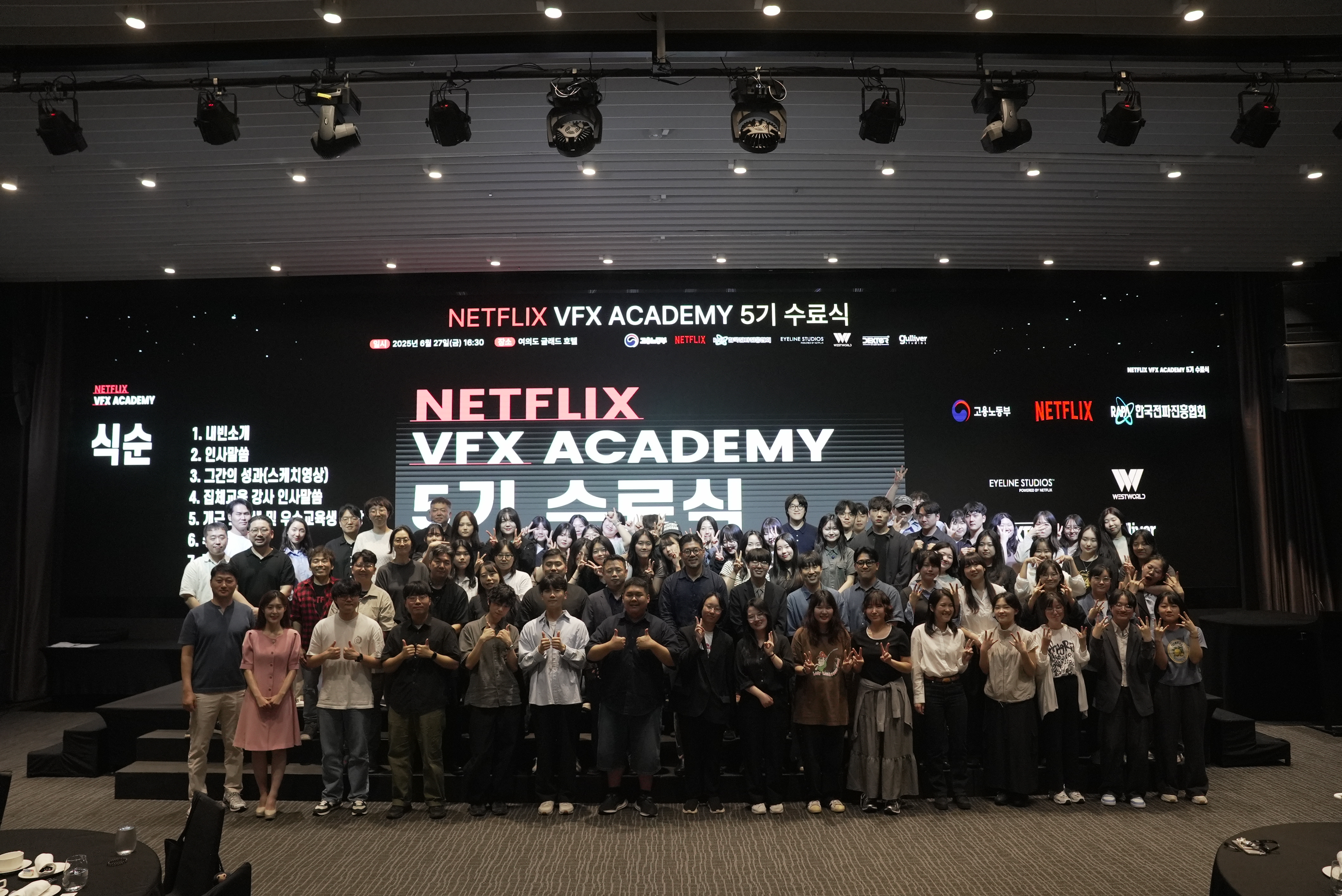 Class photo of a large group of people on risers in an auditorium giving thumbs up to the camera. Behind them, 'Netflix VFX Academy 5' is projected onto a floor-to-ceiling sized black screen.