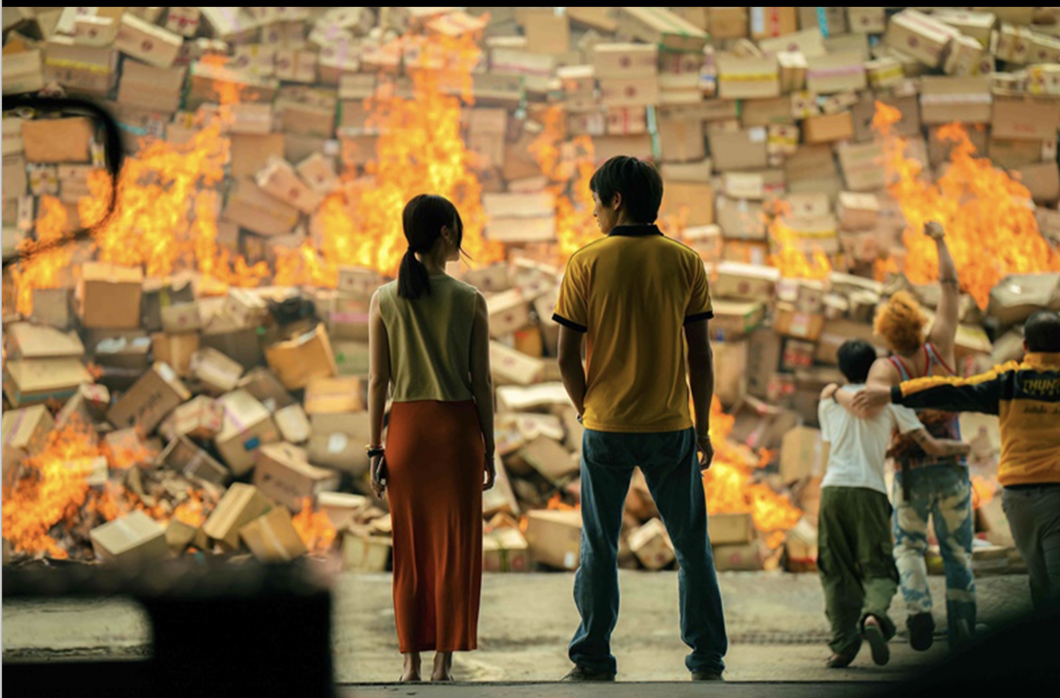 Screenshot from 'Mad Unicorn' of a man and a woman standing in front of an impossibly tall pile of packages that are all catching fire. 