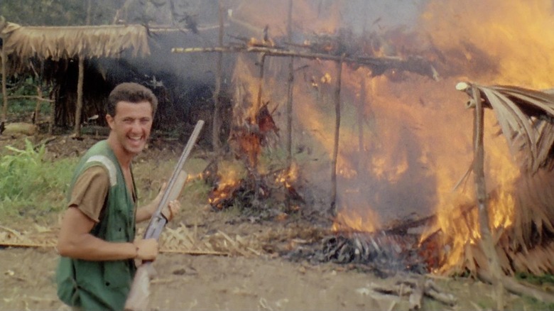 A man with a shotgun smiles near a burning thatched house in "Cannibal Holocaust" (1980)