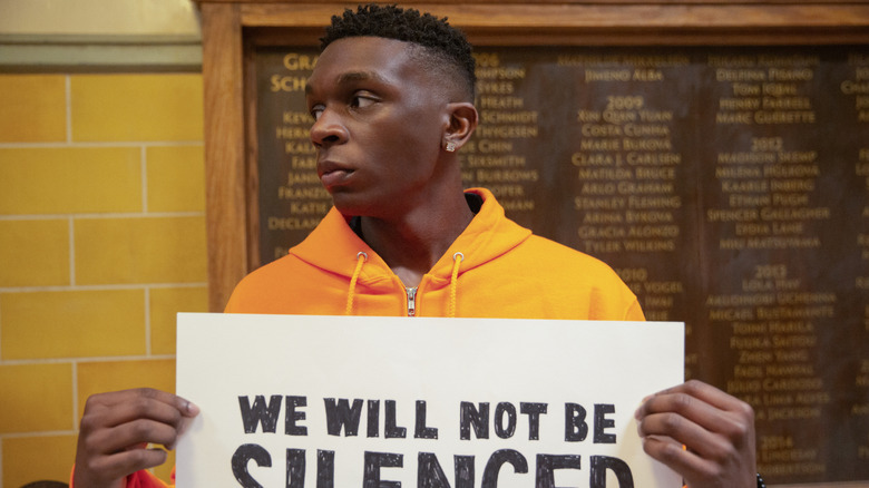 John holding a protest sign in "Grand Army."