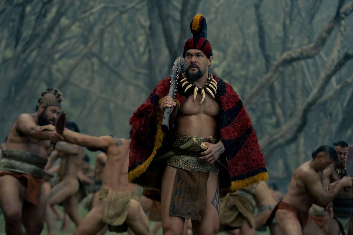 Jason Momoa in 'Chief of War,' shown here wearing a necklace of claws, a red flowery cape with yellow piping, and a red and yellow headdress to match