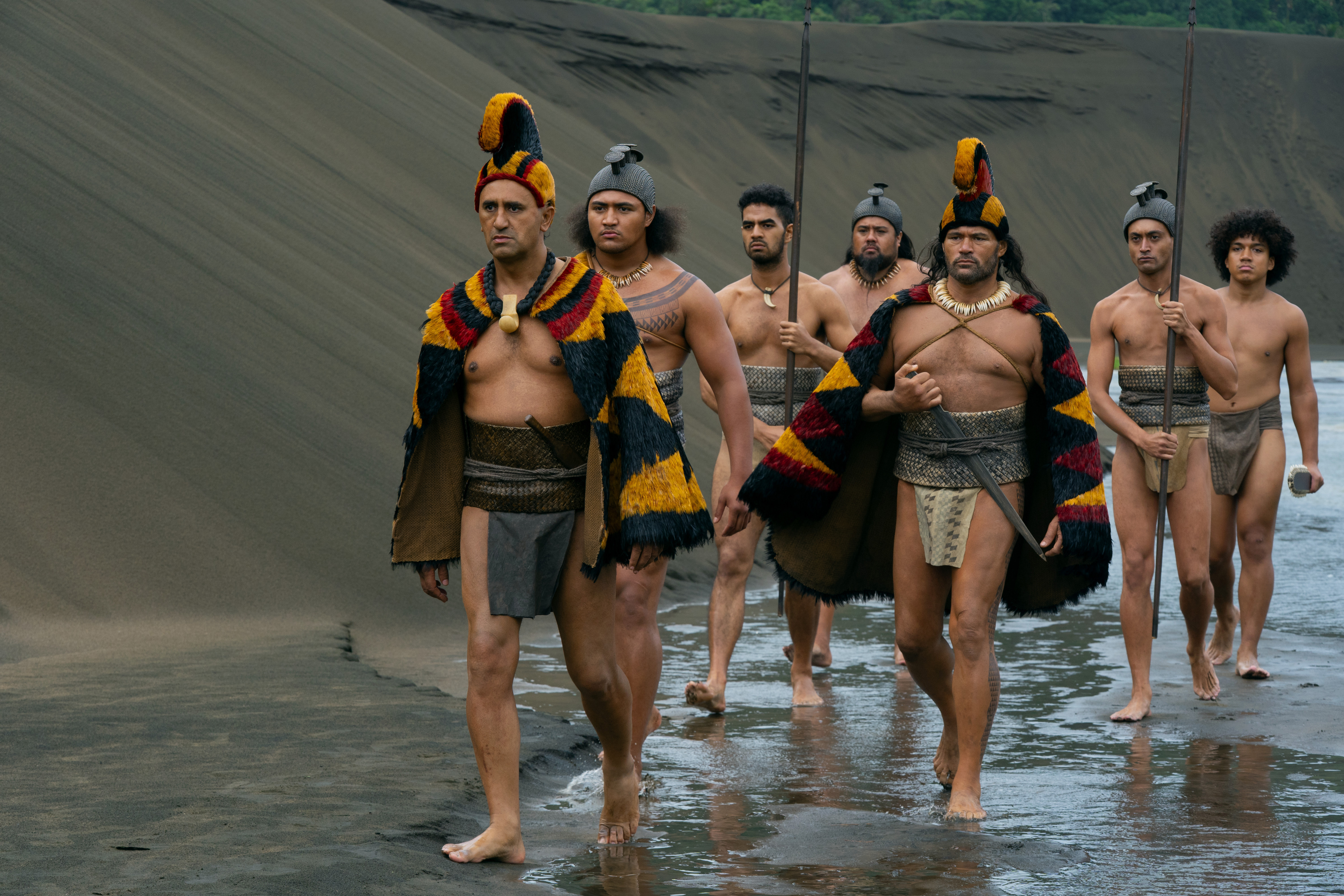 A band of Hawaiian warriors march along a beach with a dune behind them in 'Chief of War'