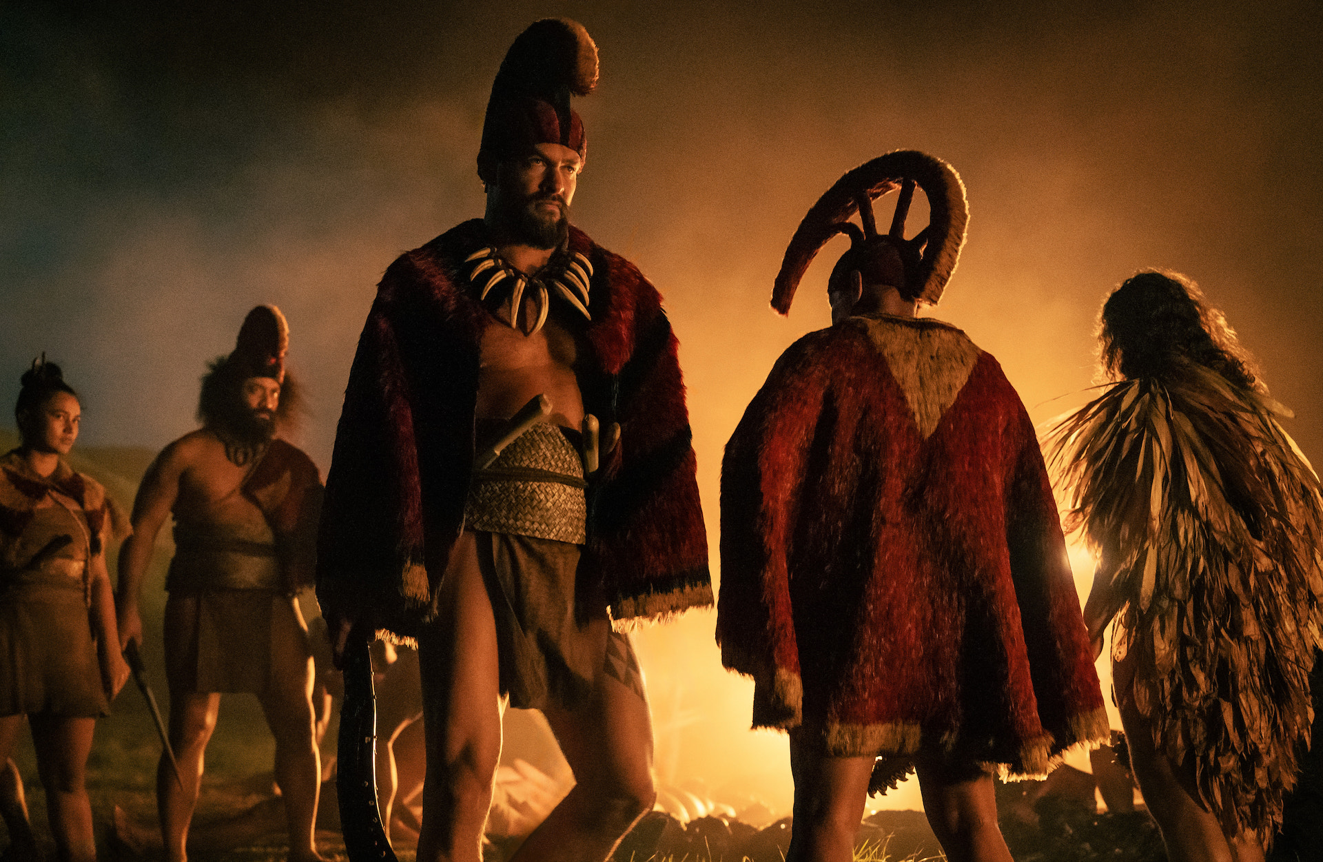 Jason Momoa standing against a firelit background in Hawaiian warrior cape and hat in 'Chief of War'