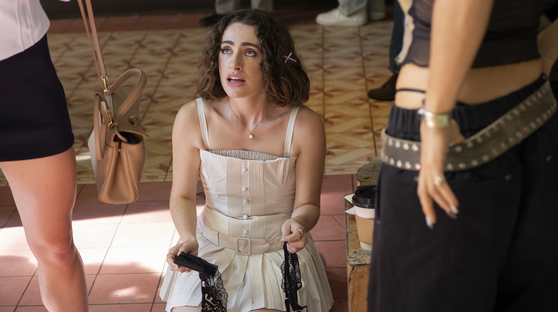A woman in a white dress crouching on the ground and holding black lace lingerie; Rachel Sennott in 'I Love LA'