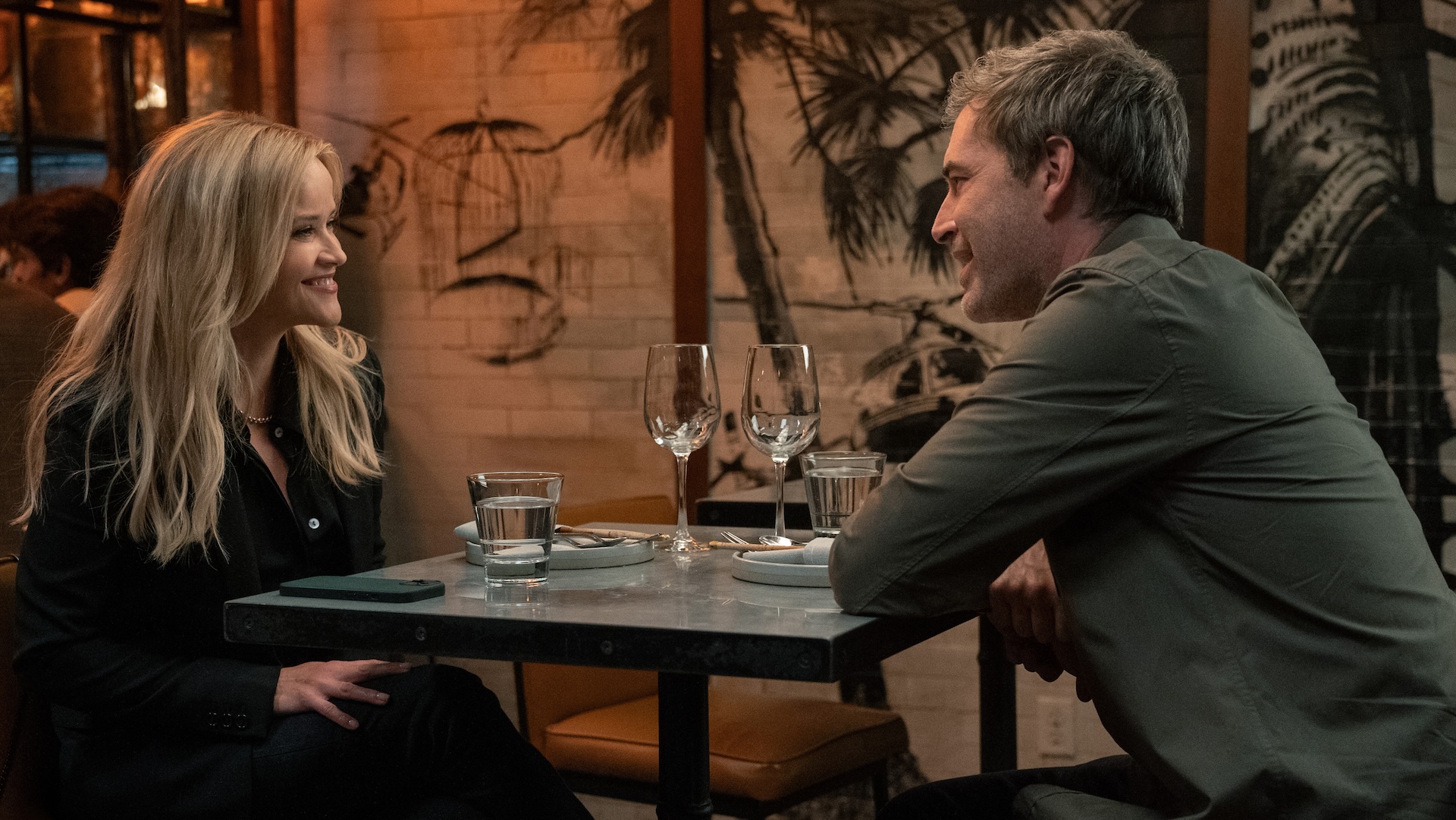 A man and woman smiling at each other across a restaurant table; Reese Witherspoon and Mark Duplass in 'The Morning Show'