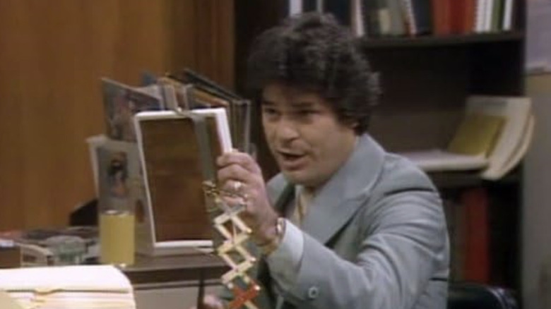Herb Tarlek at office desk holding white picture frame in left hand