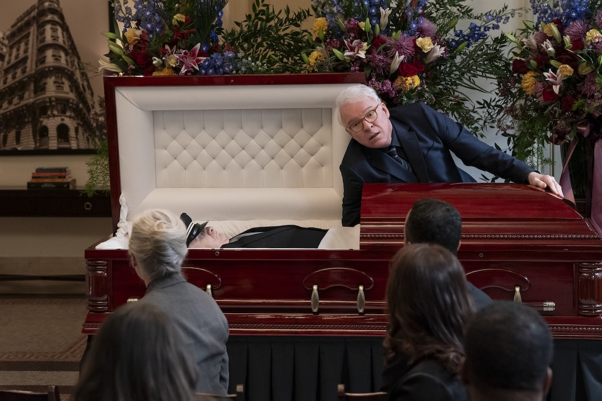 ONLY MURDERS IN THE BUILDING Season 5 stars Steve Martin as Charles Hayden Savage, shown here reaching into a coffin mid-funeral, as onlookers stare at him 