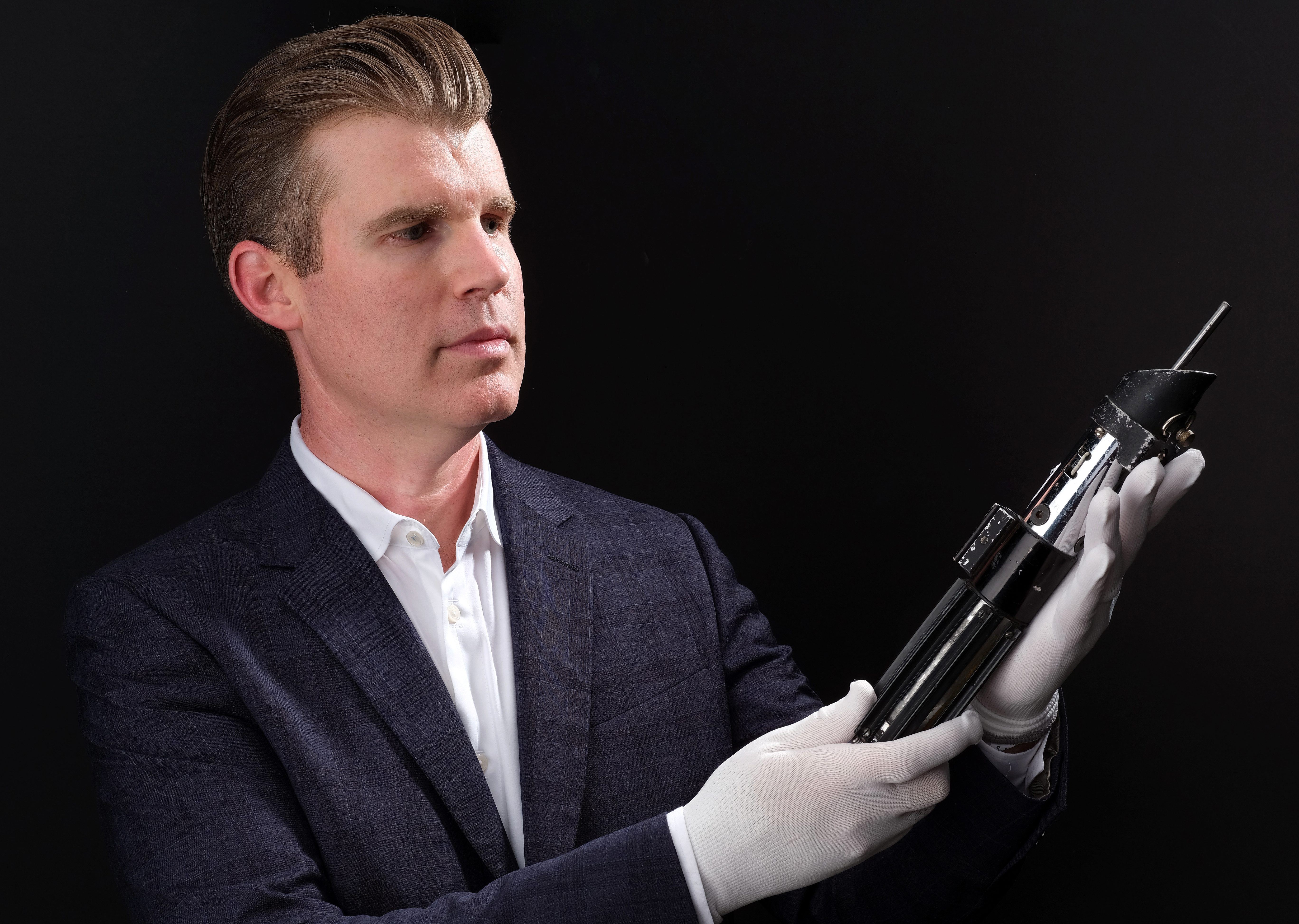 A man in a suit holding a lightsaber prop in gloved hands against a black background.