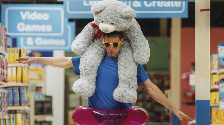 Jeff walks through Toys R Us wearing a pool floatie and a stuffed bear on his back in "Roofman" (2025)